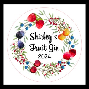 May include: A circular design featuring a watercolor wreath of green leaves, red berries, and various fruits, including strawberries, blueberries, and cherries. The text "Shirley's Fruit Gin 2024" is centered within the wreath.