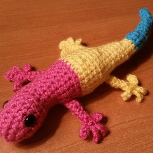May include: A crocheted lizard toy with a pink, yellow, and blue color scheme. The lizard has black eyes and a long tail.