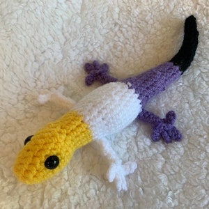 May include: A crocheted gecko plush toy with a yellow, white, and purple color scheme. The gecko has a black tail and black eyes.