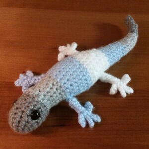 May include: A handmade crochet lizard in shades of gray, blue, and white. The lizard has a long tail, four small feet, and a detailed head with black eyes. The reptile is lying on a wooden surface.