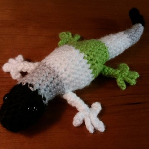 May include: A crocheted gecko plush toy with a black head, white body, and green tail. The toy is made with a soft, textured yarn.