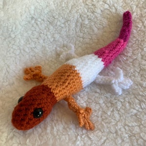May include: A crocheted gecko toy with a brown head, orange body, white middle section, and pink tail.