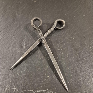 Pode incluir: Dois espetos de metal torcidos com alças arredondadas. Os espetos estão cruzados e as alças estão na parte superior da imagem.