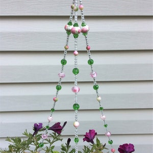 May include: A three-strand hanging planter with green and pink beads. The planter is made of clear beads with floral accents. The planter is hanging from a white ceiling.