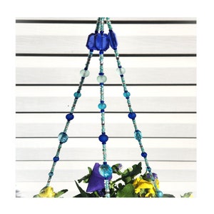 May include: A hanging planter made of blue and turquoise glass beads. The planter is suspended from a white ceiling with a plant growing inside.