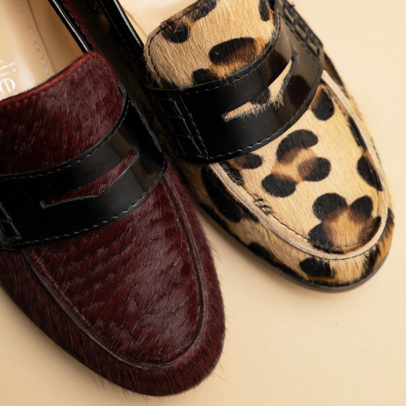 May include: Two pairs of loafers with a black leather strap across the top. One pair is burgundy with a hairy texture and the other is brown with a leopard print.