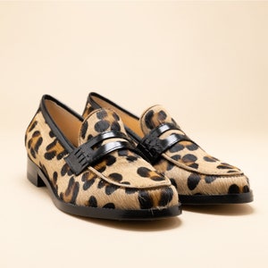 May include: A pair of leopard print loafers with black leather accents. The loafers have a classic penny loafer style with a black leather strap across the vamp.