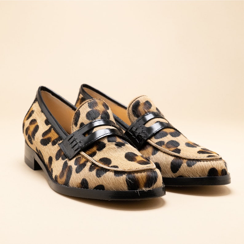May include: A pair of leopard print loafers with black leather accents. The loafers have a classic penny loafer style with a black leather strap across the vamp.