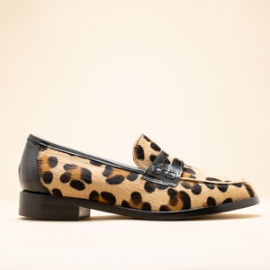 May include: A pair of leopard print loafers with black leather accents. The loafers have a classic penny loafer style with a black leather strap across the vamp.