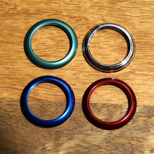 Puede incluir: Cuatro anillos metálicos de colores, verde, plateado, azul y rojo, dispuestos sobre una superficie de madera. Los anillos son circulares con un acabado liso y brillante. La imagen está bien iluminada, resaltando los colores y texturas.
