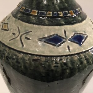 Może przedstawiać: Zielony ceramiczny wazon z białym pasem i niebieskimi i złotymi geometrycznymi wzorami. Wazon ma fakturowaną powierzchnię.