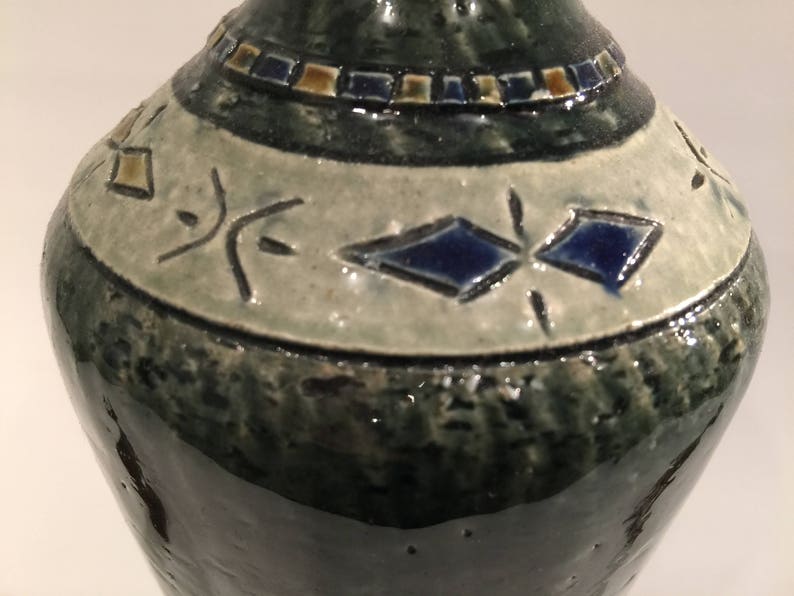 Może przedstawiać: Zielony ceramiczny wazon z białym pasem i niebieskimi i złotymi geometrycznymi wzorami. Wazon ma fakturowaną powierzchnię.