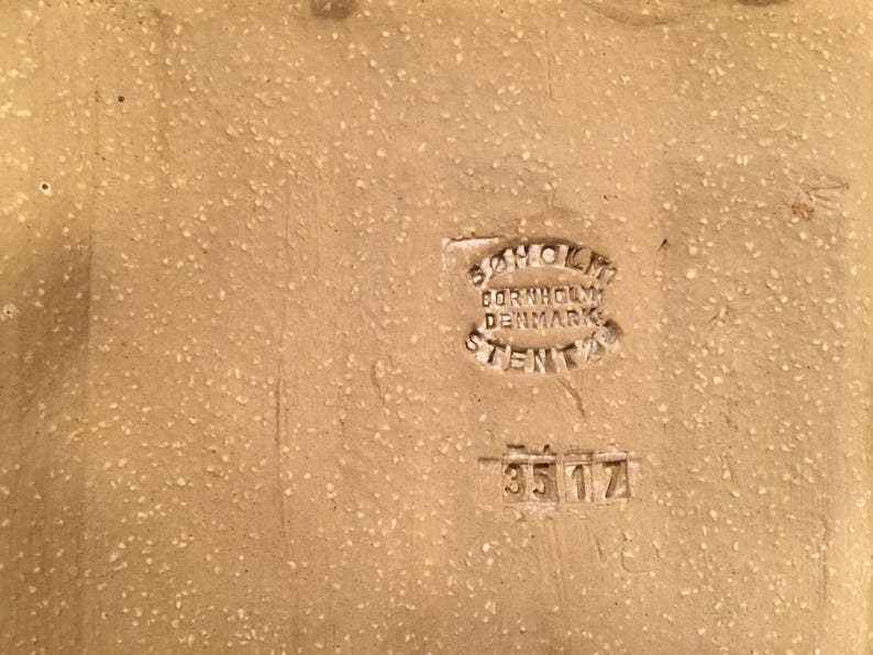 Puede incluir: Primer plano de una baldosa de cer&aacute;mica de color tostado con un sello marr&oacute;n que dice "Bornholm Denmark Stengods" y el n&uacute;mero "35 17".