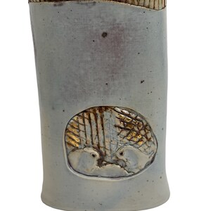 Könnte beinhalten: Eine weiße Keramikvase mit einem strukturierten Rand und einem goldenen und weißen Design von zwei Vögeln in einer runden Öffnung.