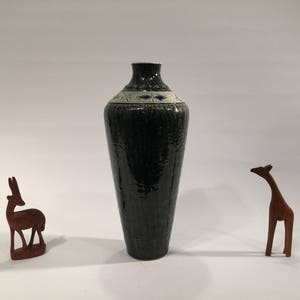 Może przedstawiać: Zielony ceramiczny wazon z białym i niebieskim paskiem z wzorem w środkowej części. Wazon jest wysoki i smukły, z zaokrąglonym dnem i wąskim otworem na g&oacute;rze.
