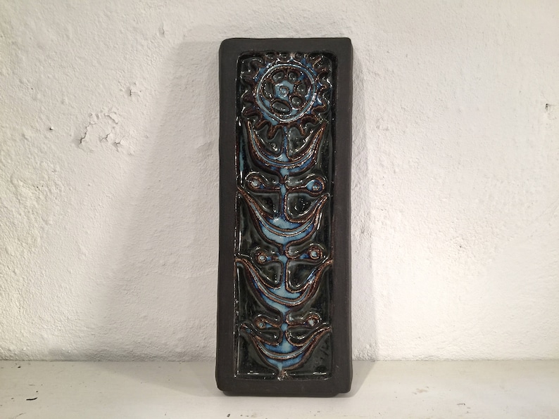 Puede incluir: Un azulejo de cer&aacute;mica marr&oacute;n oscuro con un dise&ntilde;o floral azul y blanco. El azulejo presenta una flor estilizada con un c&iacute;rculo central y cuatro p&eacute;talos. La flor est&aacute; rodeada de cuatro flores estilizadas m&aacute;s peque&ntilde;as.