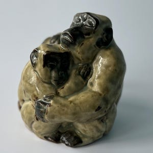 Puede incluir: Una escultura de cerámica de dos monos abrazándose. Los monos son de color marrón claro con detalles en marrón oscuro. La escultura es una representación realista de los animales.