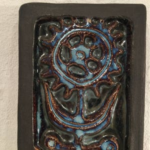 Puede incluir: Un azulejo de cer&aacute;mica azul y marr&oacute;n con un dise&ntilde;o de flor estilizado. El azulejo est&aacute; enmarcado en un borde negro.