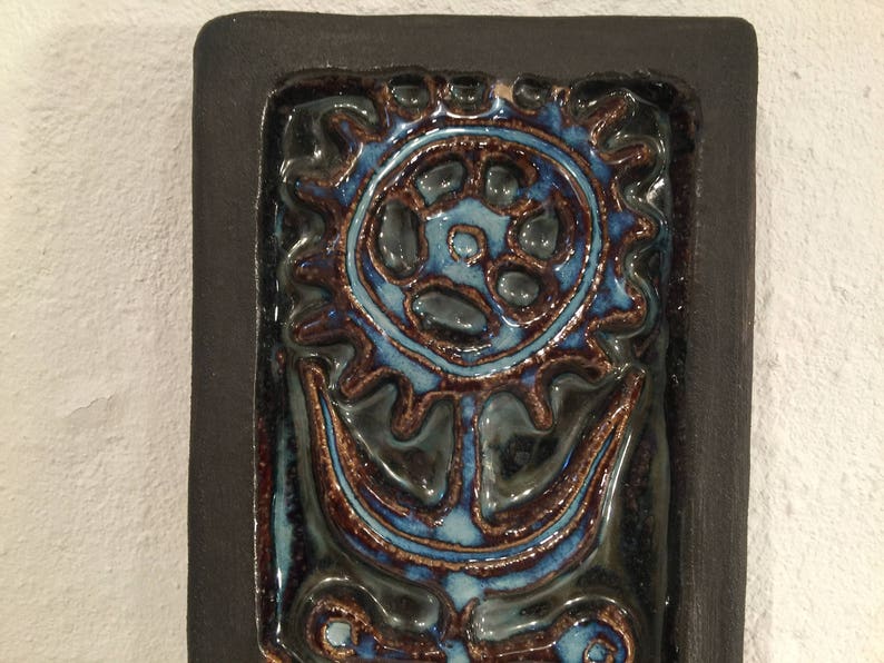 Puede incluir: Un azulejo de cer&aacute;mica azul y marr&oacute;n con un dise&ntilde;o de flor estilizado. El azulejo est&aacute; enmarcado en un borde negro.