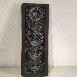 Puede incluir: Un azulejo de pared de cer&aacute;mica con un marco marr&oacute;n oscuro y un dise&ntilde;o floral azul y marr&oacute;n. El azulejo presenta una flor estilizada con un c&iacute;rculo central y cuatro p&eacute;talos.