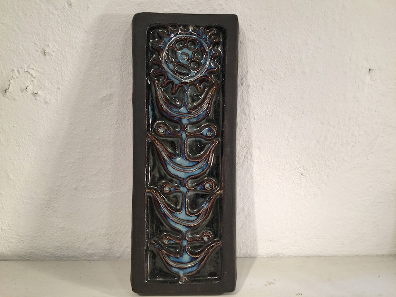 Puede incluir: Un azulejo de pared de cer&aacute;mica con un marco marr&oacute;n oscuro y un dise&ntilde;o floral azul y marr&oacute;n. El azulejo presenta una flor estilizada con un c&iacute;rculo central y cuatro p&eacute;talos.