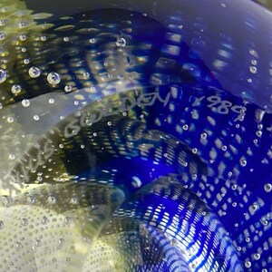K&ouml;nnte beinhalten: Ein blauer Glas-Briefbeschwerer mit einer wei&szlig;en Signatur, die "Nils Goebel 1984" lautet, und einem wei&szlig;en Muster aus Blasen.