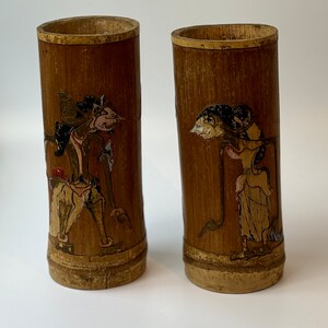 Può includere: Due tazze di bambù marroni con figure dipinte in oro, rosso, nero e bianco. Le figure sono stilizzate e sembrano provenire da uno stile d'arte asiatico tradizionale.