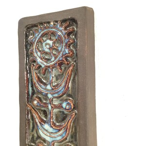 Puede incluir: Un azulejo de pared de cer&aacute;mica marr&oacute;n con un dise&ntilde;o floral estilizado azul y marr&oacute;n. El azulejo tiene una superficie texturizada en relieve.