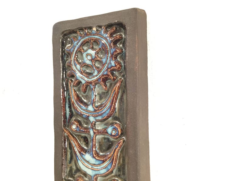 Puede incluir: Un azulejo de pared de cer&aacute;mica marr&oacute;n con un dise&ntilde;o floral estilizado azul y marr&oacute;n. El azulejo tiene una superficie texturizada en relieve.