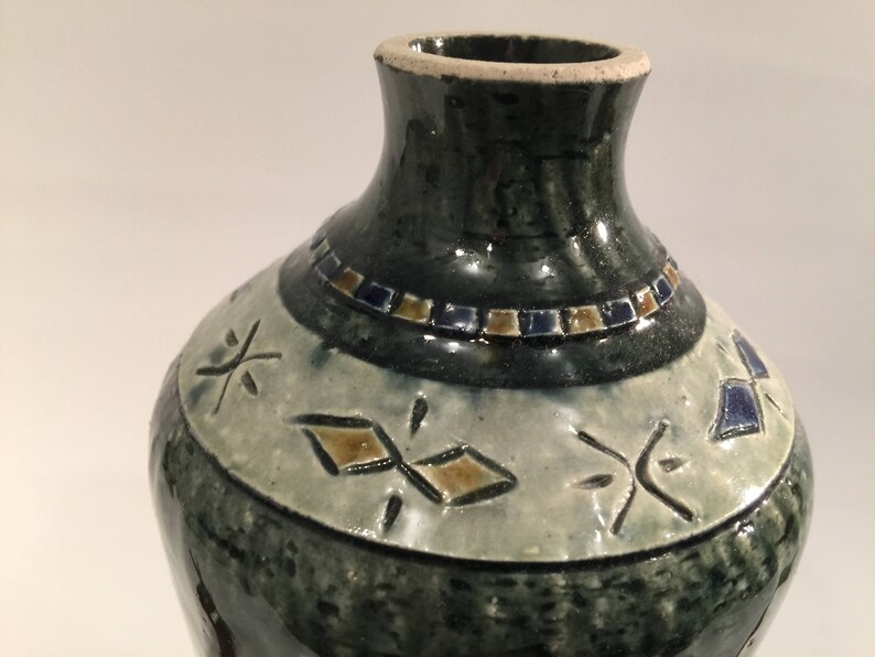 Może przedstawiać: Zielony ceramiczny wazon z białym pasem wok&oacute;ł środka. Pas ma geometryczny wz&oacute;r w kolorze brązowym i niebieskim. Wazon ma wąską szyjkę i szeroką podstawę.