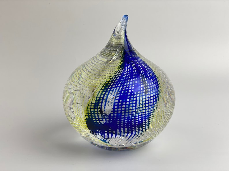 K&ouml;nnte beinhalten: Ein durchsichtiger Glas-Briefbeschwerer mit einem geometrischen Muster in Blau und Gelb. Das Glas ist mit kleinen Blasen strukturiert.
