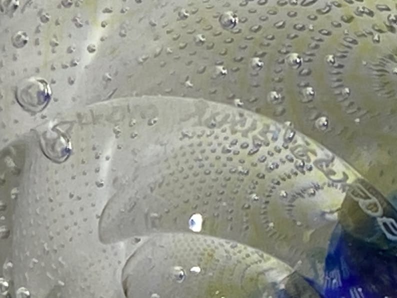 K&ouml;nnte beinhalten: Nahaufnahme eines durchsichtigen Glas-Papiergewichts mit einer strukturierten Oberfl&auml;che und kleinen Luftblasen, die darin eingeschlossen sind. Das Glas hat ein blaues und wei&szlig;es Spiralmuster.