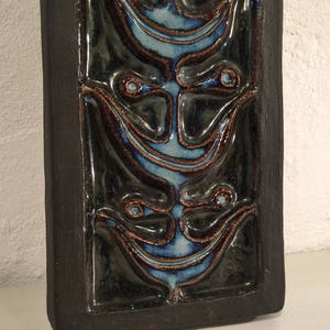 Puede incluir: Un azulejo de cer&aacute;mica marr&oacute;n oscuro con un dise&ntilde;o en relieve de tres caras estilizadas. Las caras est&aacute;n delineadas en azul y tienen un patr&oacute;n de remolino.