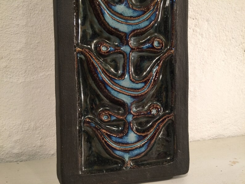 Puede incluir: Un azulejo de cer&aacute;mica marr&oacute;n oscuro con un dise&ntilde;o en relieve de tres caras estilizadas. Las caras est&aacute;n delineadas en azul y tienen un patr&oacute;n de remolino.