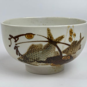 May include: A white ceramic bowl with a brown and gold fish design. The bowl has a rounded shape and a small foot.