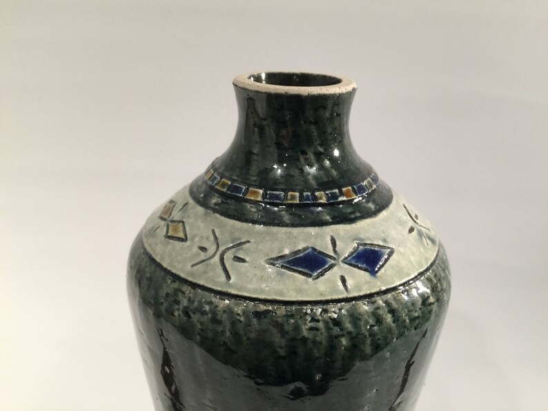 Może przedstawiać: Zielony ceramiczny wazon z białym paskiem i niebieskimi i złotymi geometrycznymi wzorami.