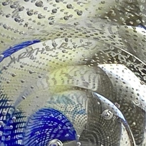 K&ouml;nnte beinhalten: Nahaufnahme eines durchsichtigen Glas-Papiergewichtes mit einem blauen und wei&szlig;en Muster und kleinen Luftblasen. Das Papiergewicht tr&auml;gt die Aufschrift "X-Ray" auf der Seite.