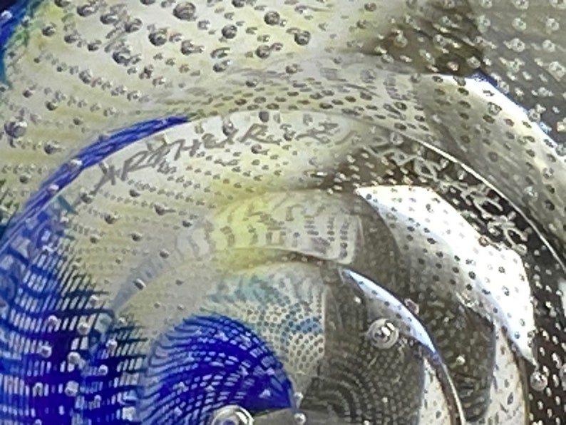 K&ouml;nnte beinhalten: Nahaufnahme eines durchsichtigen Glas-Papiergewichtes mit einem blauen und wei&szlig;en Muster und kleinen Luftblasen. Das Papiergewicht tr&auml;gt die Aufschrift "X-Ray" auf der Seite.