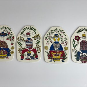 Puede incluir: Cuatro tablas de cortar de cerámica con diseños coloridos de arte popular que representan personas con ropa tradicional y motivos florales. Las tablas son blancas con acentos rojos, azules, amarillos y verdes.