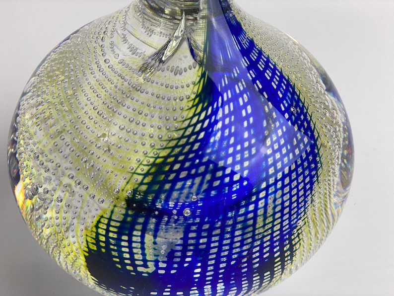K&ouml;nnte beinhalten: Eine klare Glasvase mit einem geometrischen Muster in Blau, Gr&uuml;n und Gelb. Die Vase hat eine strukturierte Oberfl&auml;che mit kleinen Blasen.