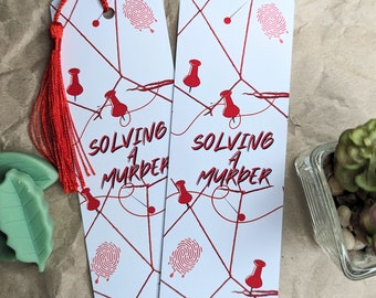 May include: Two white bookmark with red string and red push pins. The bookmarks have the text "Solving a Murder" on them.