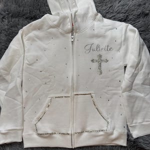 Könnte beinhalten: Weißer Kapuzenpullover mit Reißverschluss, einem Kreuz und dem Namen Juliette in Strasssteinen.