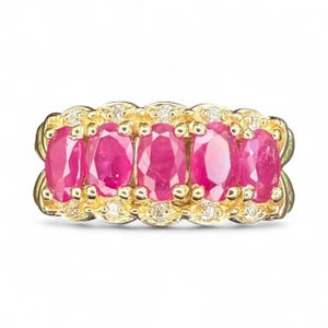 May include: A gold ring featuring five oval-cut, vibrant pink rubies. The rubies are set in a row, surrounded by smaller diamonds and a decorative gold setting. A luxurious piece of jewelry.