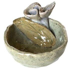 May include: A small, light green ceramic bowl with a textured surface. A light purple and brown ceramic flower sits on top of the bowl. The flower has a leaf-like shape.