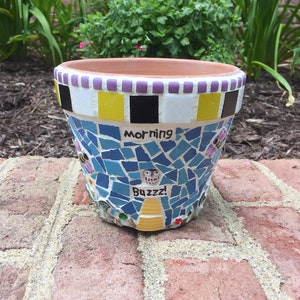 May include: A blue and yellow mosaic flower pot with a white rim and a black and white checkered pattern. The pot has the words "Morning Buzzzz!" written on it.