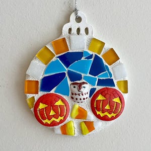 May include: A round, mosaic-style Halloween ornament. The ornament features a white base with colorful glass tiles in shades of blue, orange, yellow, and white. It includes two red jack-o'-lanterns, a small owl, and candy corn accents. A silver string is attached for hanging.