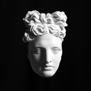 Puede incluir: Un busto de yeso blanco de una mujer con un peinado detallado. El busto está sobre un fondo negro.