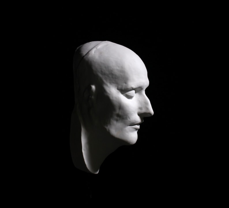 May include: A white plaster cast of a person's face, showing the profile view of the head and neck. The face is expressionless, with closed eyes and a slightly parted mouth.