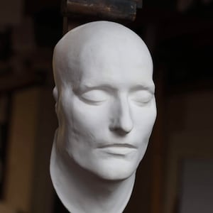 May include: A white plaster cast of a person's face with eyes closed. The cast is mounted on a wooden easel.