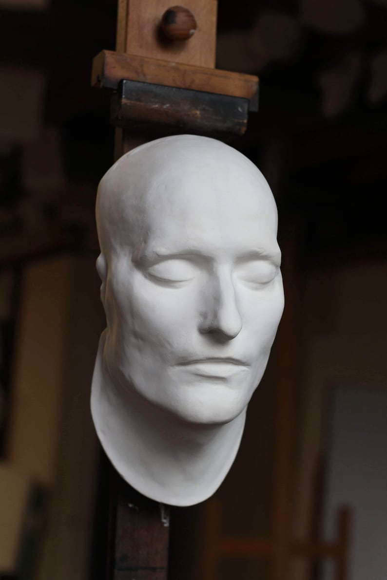 May include: A white plaster cast of a person's face with eyes closed. The cast is mounted on a wooden easel.